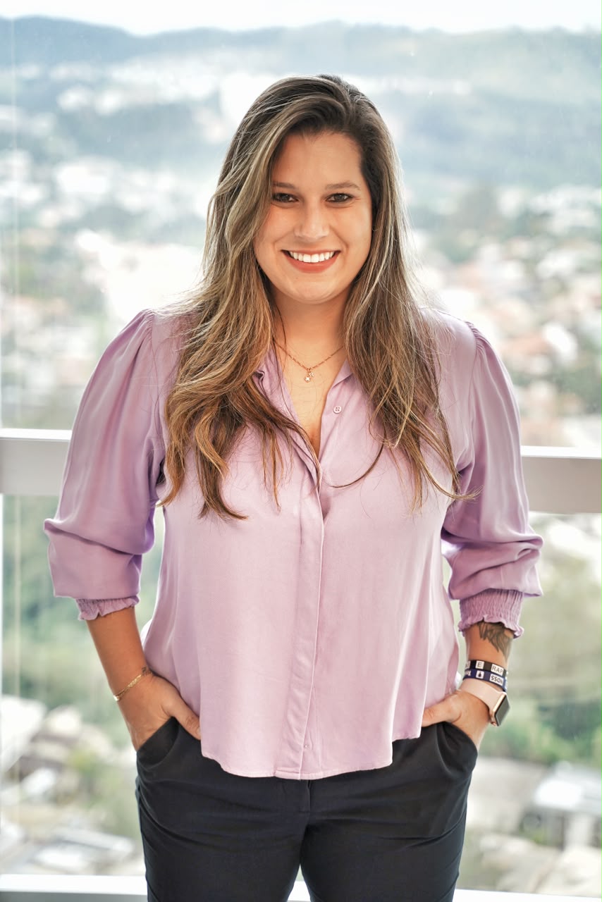 Naya sorrindo, camisa lilás, com vista da cidade ao fundo